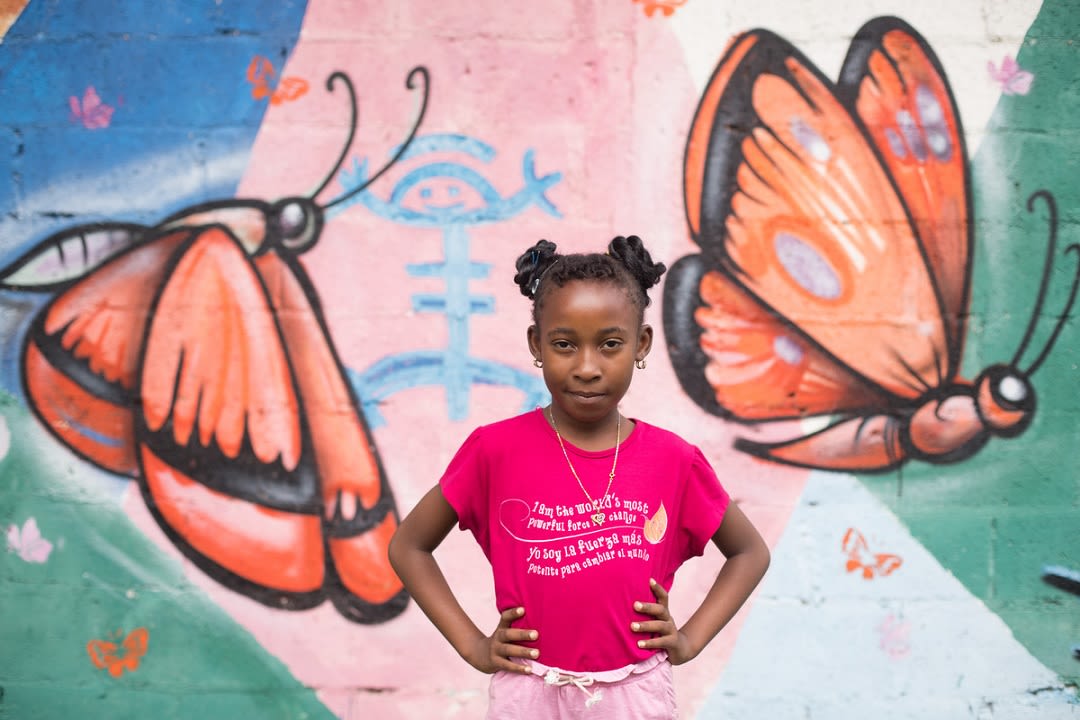 10-year-old Yohanna stands with her hands on her hips, in front of a mural of a butterfly. Her t-shirt says "I am the world's most powerful force for change."
